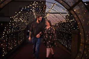 A couple walks through an illuminated tunnel archway decorated with warm glowing string lights at night.