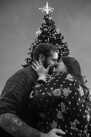 A romantic couple shares an intimate moment kissing in front of a decorated Christmas tree in black and white.
