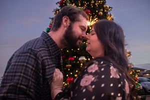 A romantic couple shares a tender kiss in front of a Christmas tree with twinkling lights in the background.