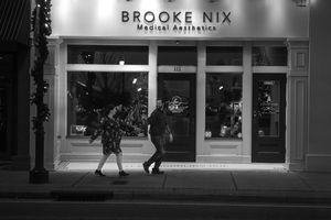 Two silhouettes walk past illuminated Brooke Nix storefront at night in black and white photography.