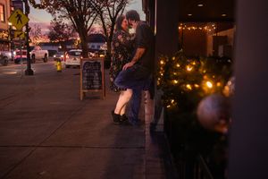 A couple embraces on a city sidewalk at dusk with holiday lights glowing in the background.