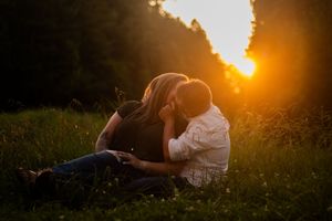Two people share an intimate moment in a meadow at sunset as golden light streams through trees in the background.