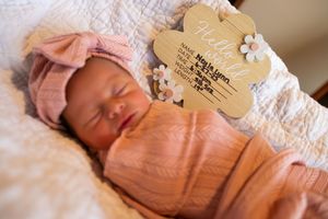Newborn baby wearing pink sleeper outfit rests peacefully on white blanket with wooden heart decoration nearby.