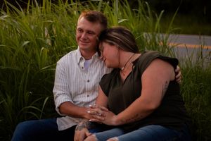 A couple sits together outdoors in tall grass during golden hour, sharing an intimate moment.
