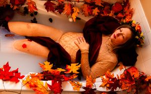 Person lying on wooden floor surrounded by scattered autumn maple leaves in warm lighting.