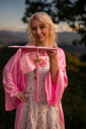 Graduate holding pink tassel and wearing pink robe against sunset mountain backdrop.