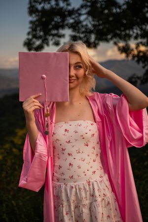 A person in a floral dress and pink cardigan poses with a book against a sunset backdrop.