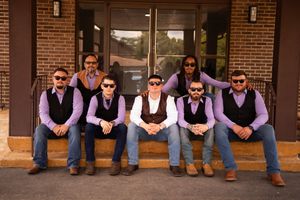 Groomsmen in matching purple vests and jeans sit casually on steps outside a brick building.