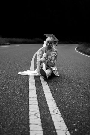 Graduate sitting on road with white lines wearing cap and gown in black and white.