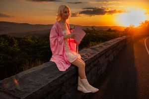 Graduate in pink robe sits on stone wall reading during golden sunset.