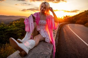 Graduate in pink robe and white boots sits on roadside barrier at sunset.