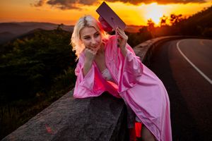 Graduate poses with diploma on mountain road barrier during dramatic sunset.
