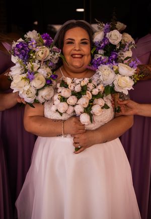 A joyful bride in a white dress holds a stunning bouquet of purple and white flowers while wearing pearl accessories.
