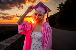 Graduate in pink robe and white dress adjusts cap against dramatic sunset sky.