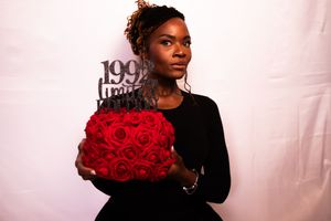 A person in a black sweater poses with a vibrant bouquet of red roses against a white backdrop.