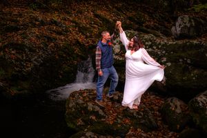 A couple in wedding attire poses among autumn leaves and trees in a romantic outdoor forest setting.
