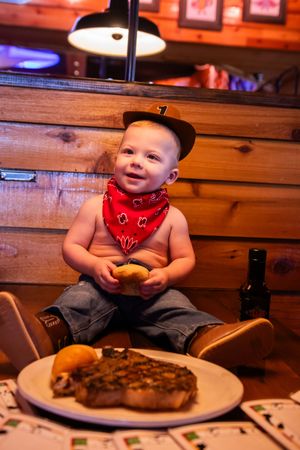 A toddler in a red bandana bib sits at a rustic wooden table in a restaurant with plates of food.