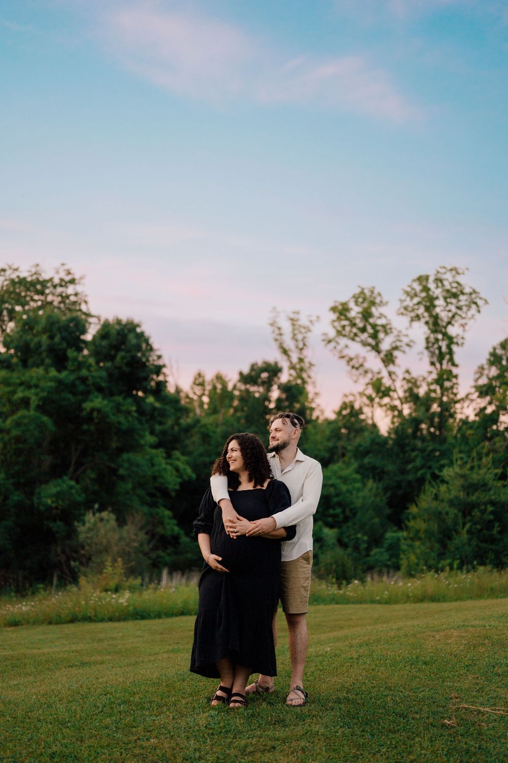 Couple embracing outdoors at sunset, pregnant woman in black dress, man in white shirt, lush green trees background.