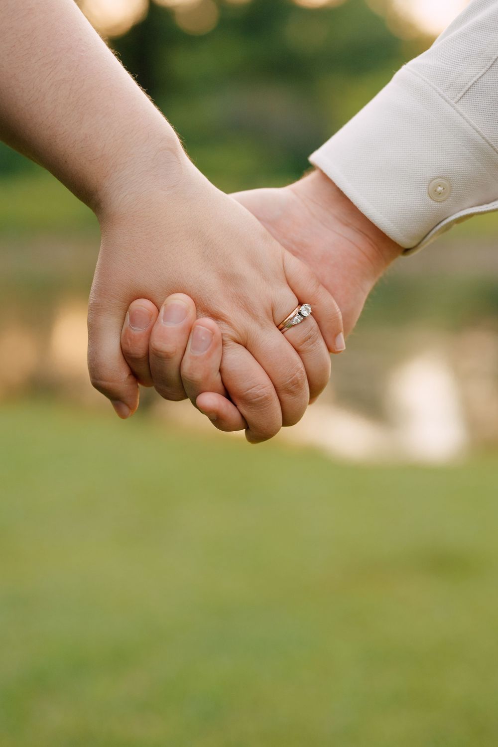 Couple holding hands outdoors with engagement ring visible, romantic close-up shot on green background.