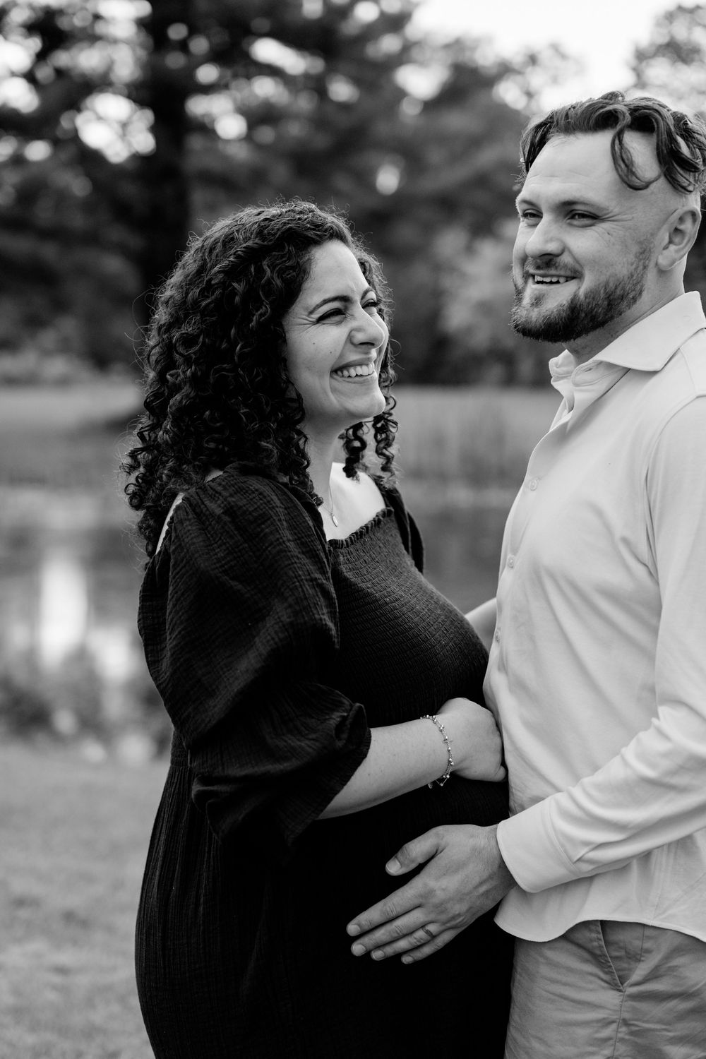 Black and white photo of a smiling pregnant woman and man tenderly cradling her baby bump outdoors near a pond.