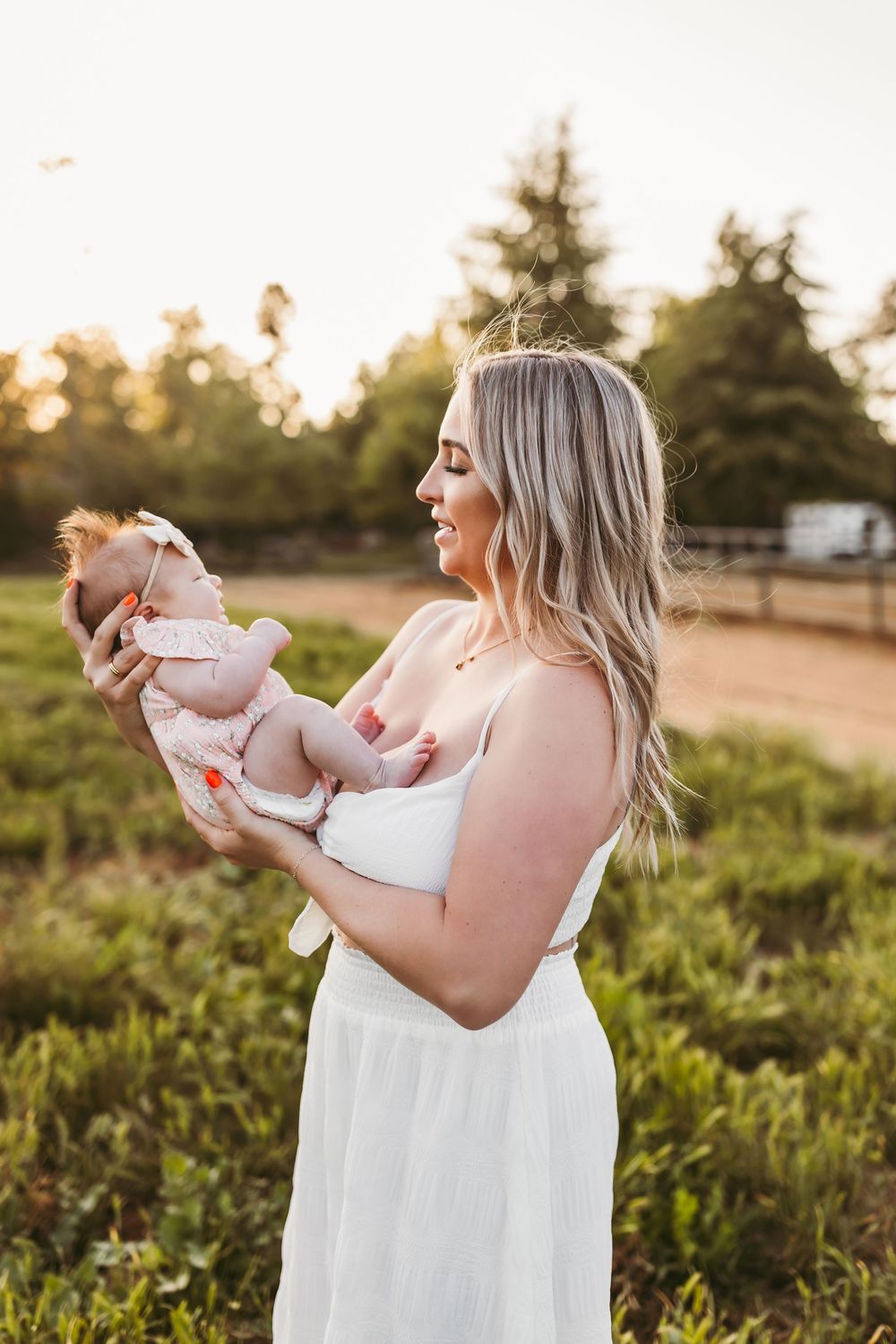 A woman in a white dress holds a baby during golden hour in a grassy field at sunset.