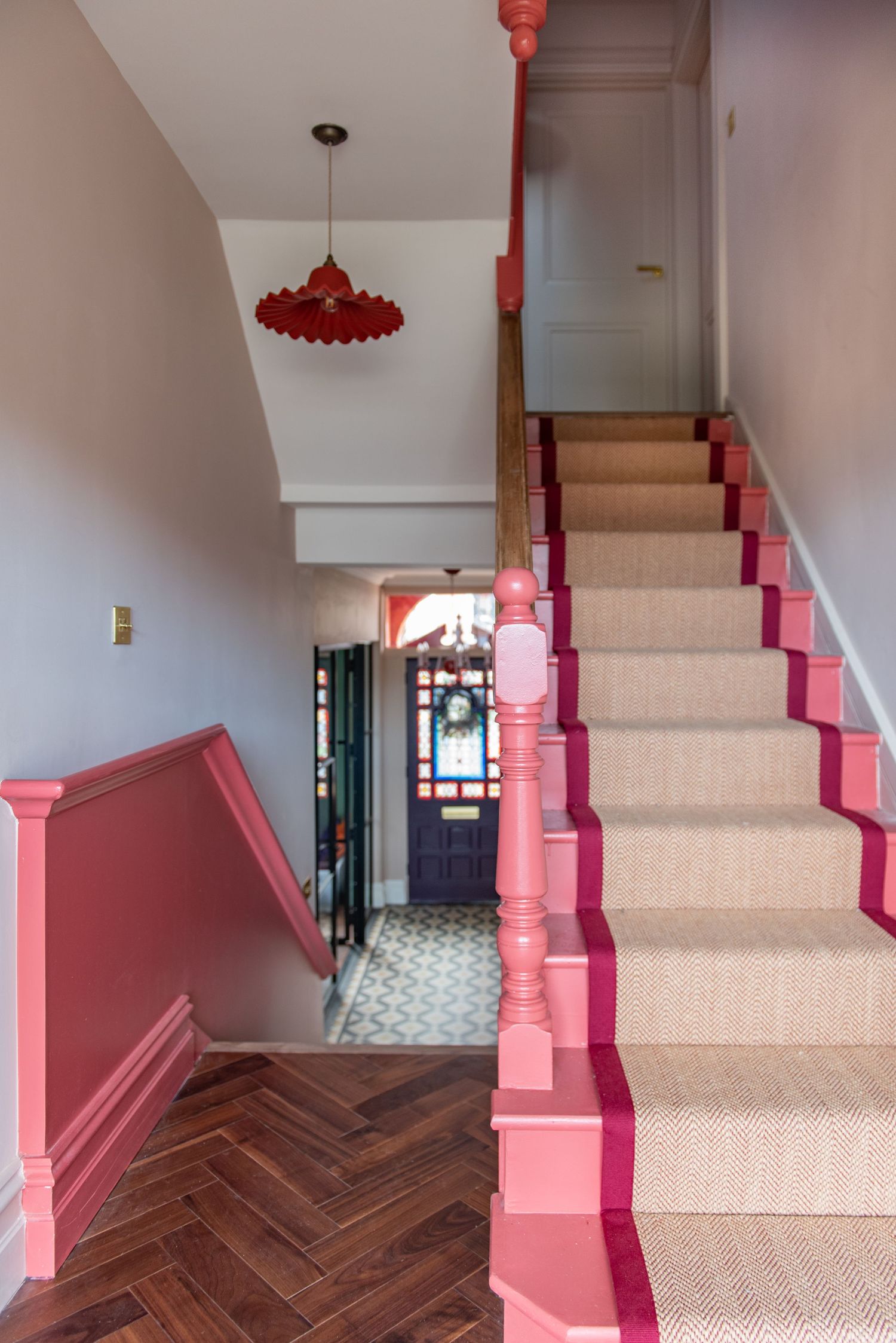 Interior staircase with pink painted railings, beige carpet runner, herringbone wood floors and a red pendant light fixture.