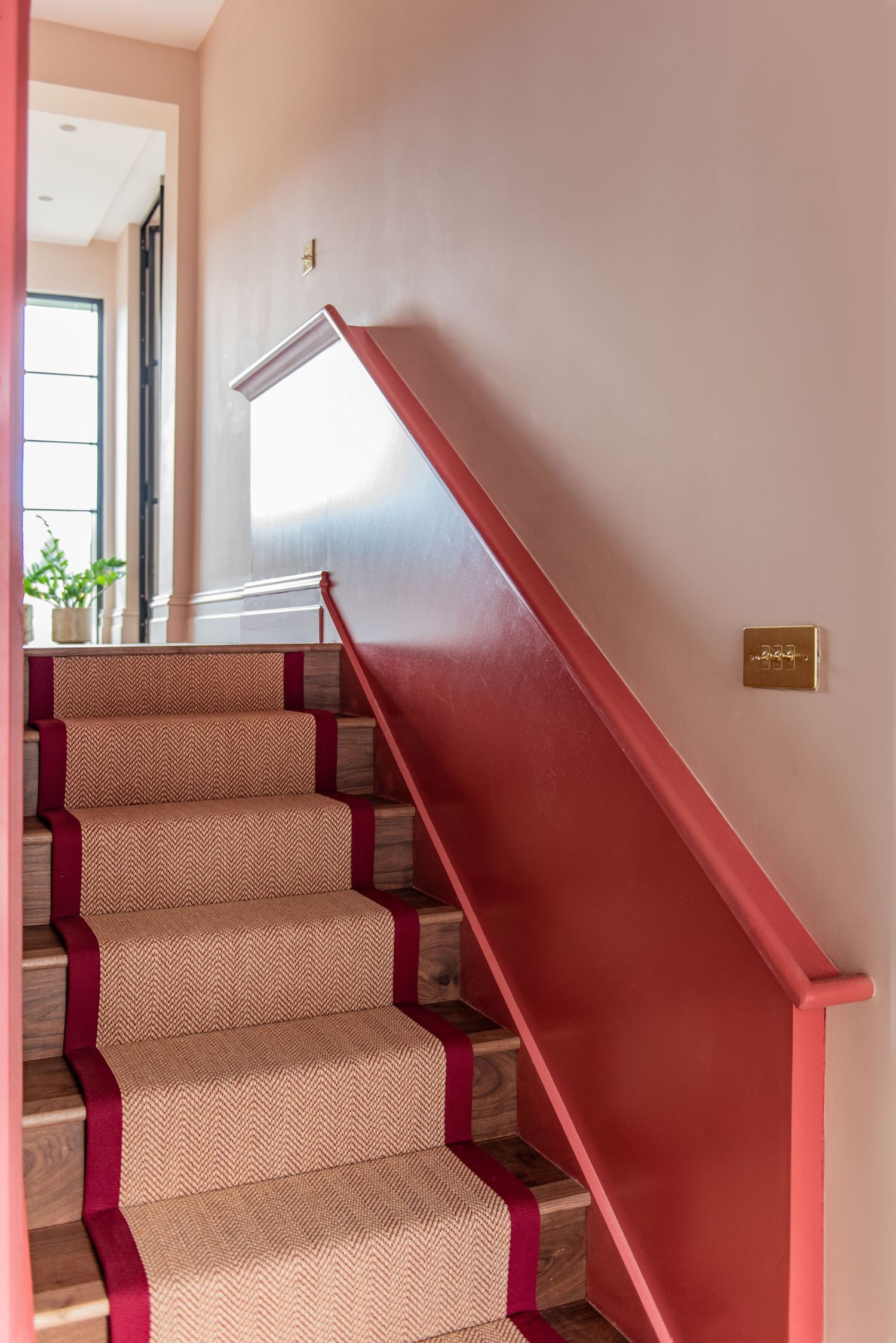 Interior staircase with beige carpet runner bordered in burgundy and matching painted handrail in residential hallway.