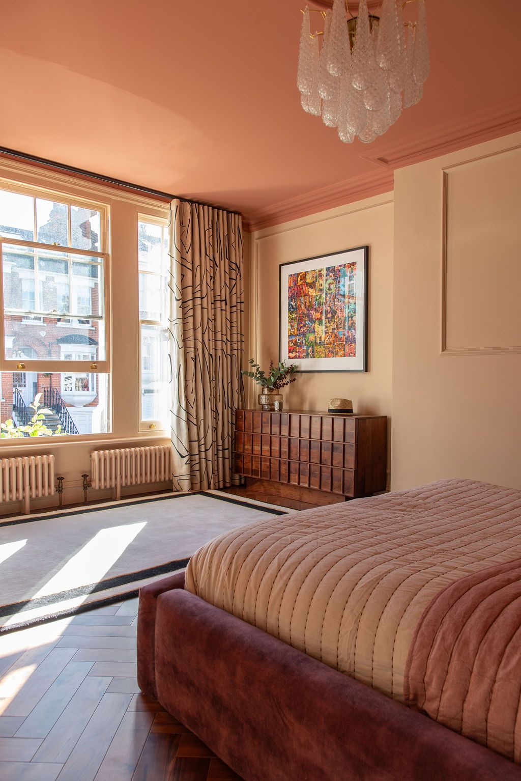 A panoramic view of a modern bedroom with cream walls, chandelier, and large windows letting in natural sunlight. Terracotta ceiling