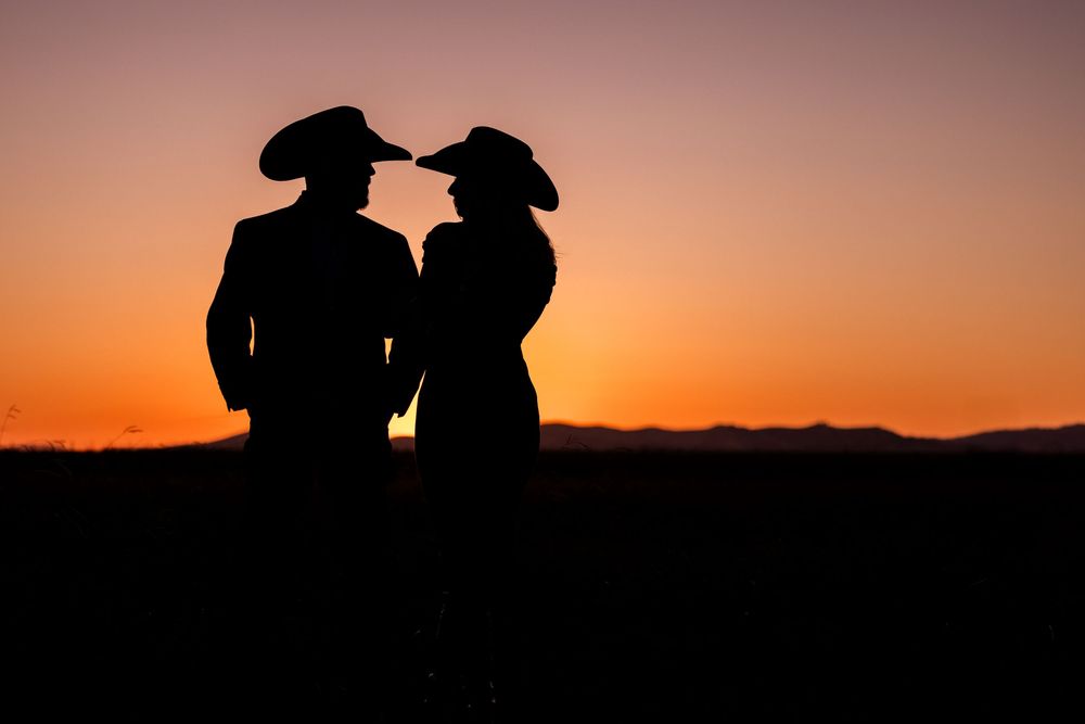 Silhouette of two people in cowboy hats standing close together against a vibrant orange sunset sky.