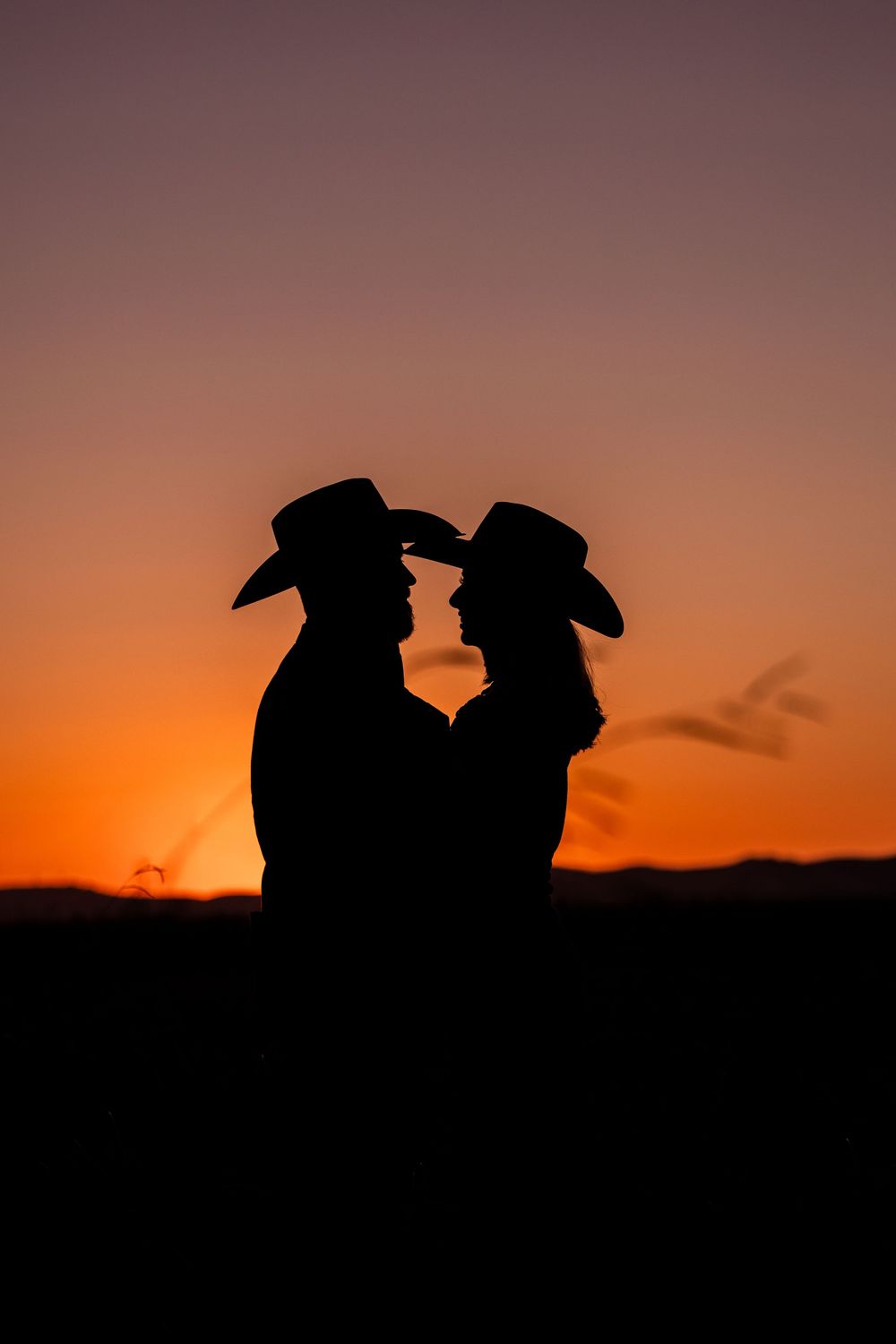 Two silhouetted figures wearing cowboy hats stand against a vibrant orange sunset sky in a dramatic western scene.