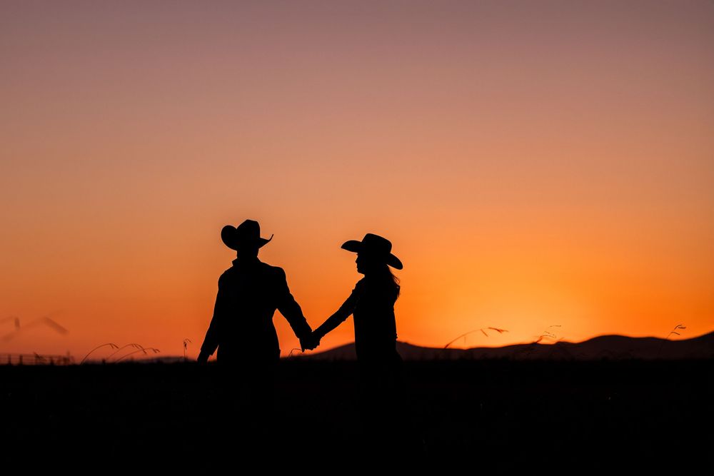 Silhouettes of people in cowboy hats against a vibrant orange sunset over mountains create a western scene.