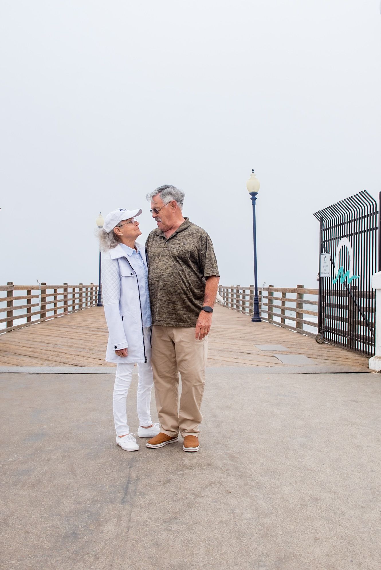 Two people in casual attire stand together on a concrete pier with a wooden boardwalk and metal railing visible.