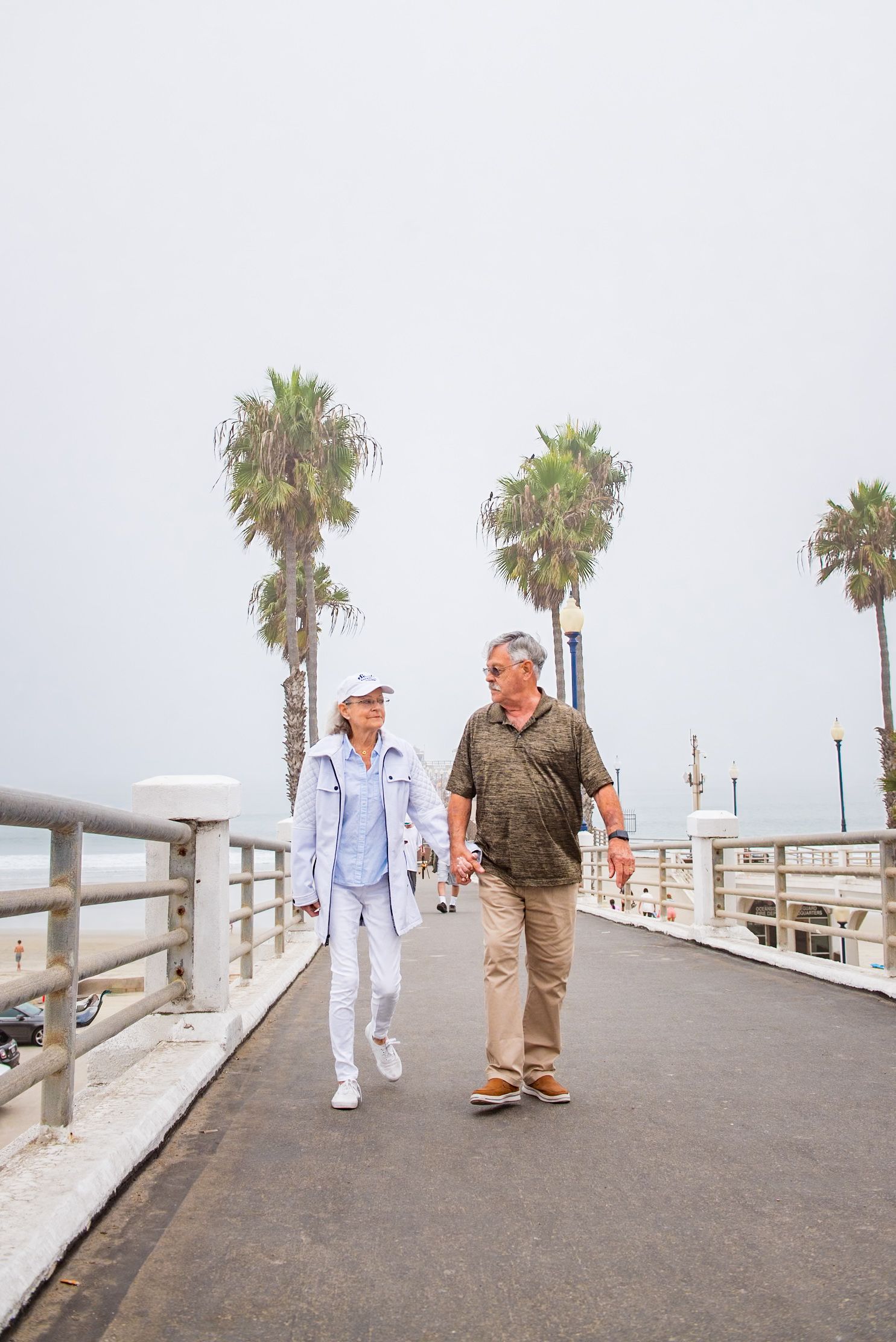 A couple takes a romantic stroll along a palm-lined pier walkway on an overcast day.