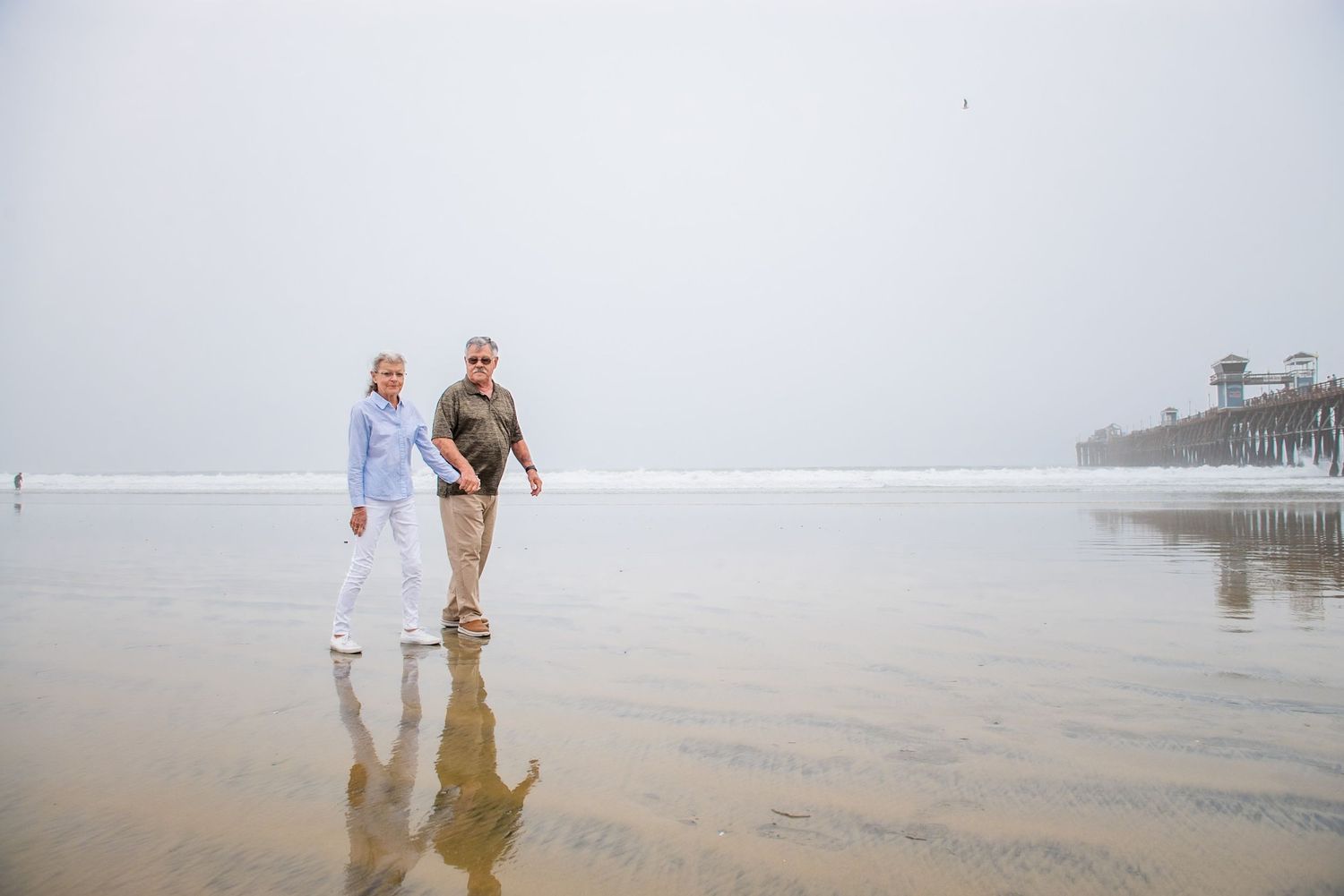 Couple walking along misty beach shore holding hands in soft morning light.