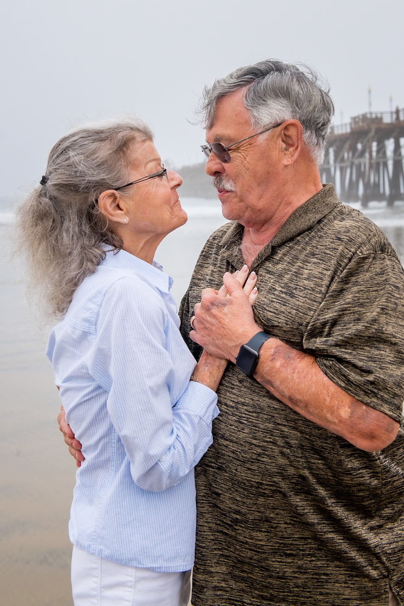 A tender moment captured between an elderly couple embracing on a misty beach with a pier in the background.