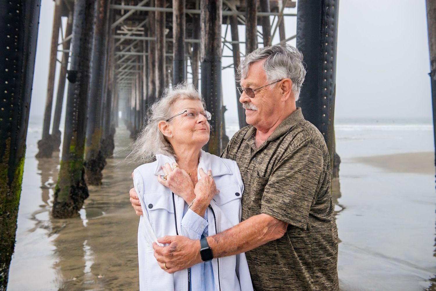 An elderly couple embraces on a beach pier while waves crash beneath them during a cloudy day.