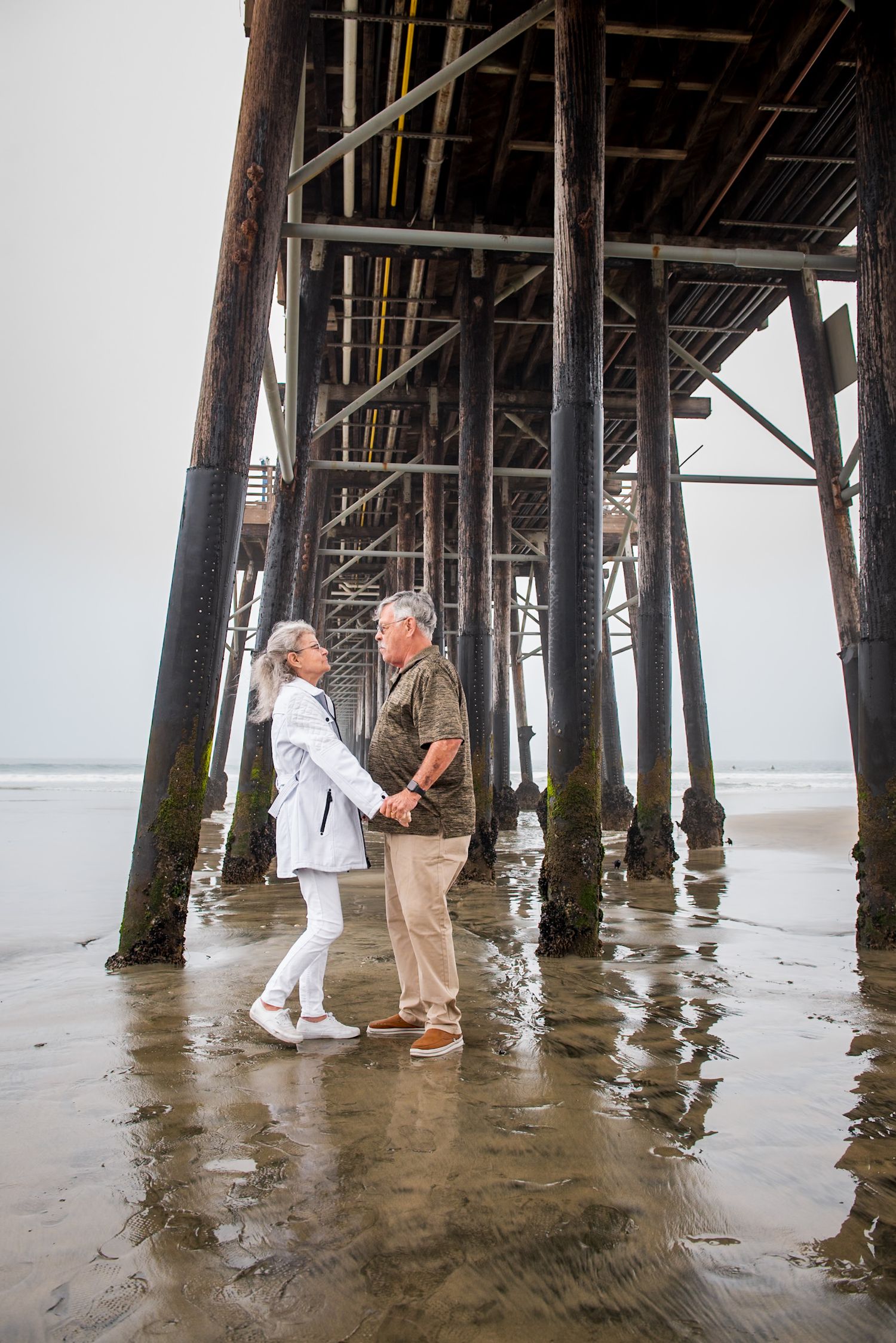 A couple shares a tender moment under a wooden pier at the beach during low tide.