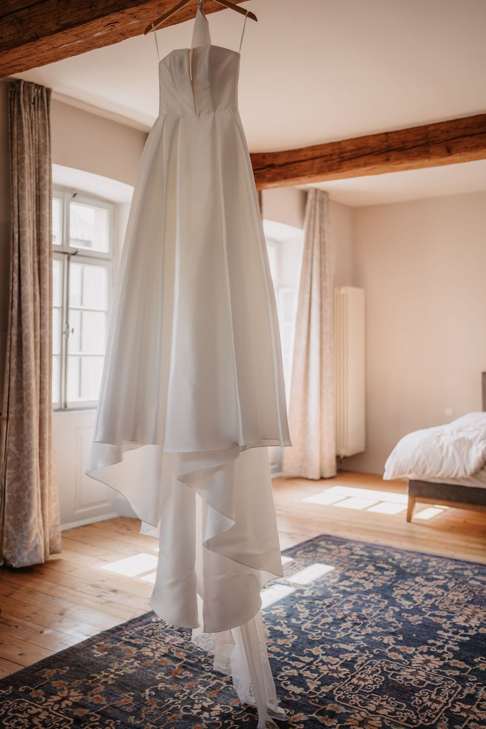 An elegant white wedding dress hangs from exposed wooden beams in a rustic bedroom with hardwood floors and an ornate rug.