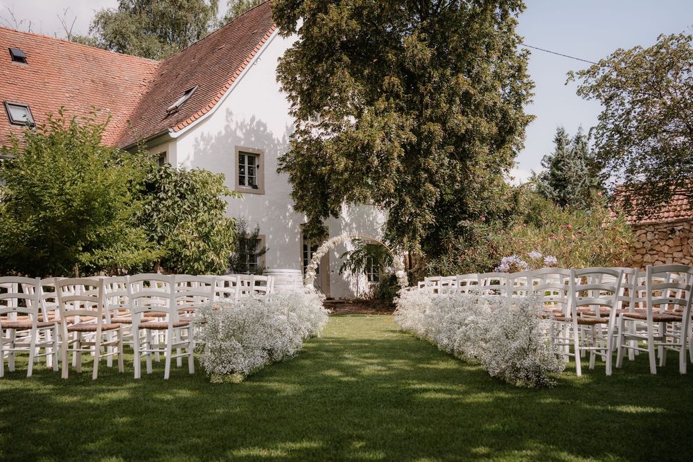 White wooden chairs arranged in rows on a green lawn for an outdoor wedding ceremony at a rustic white building.