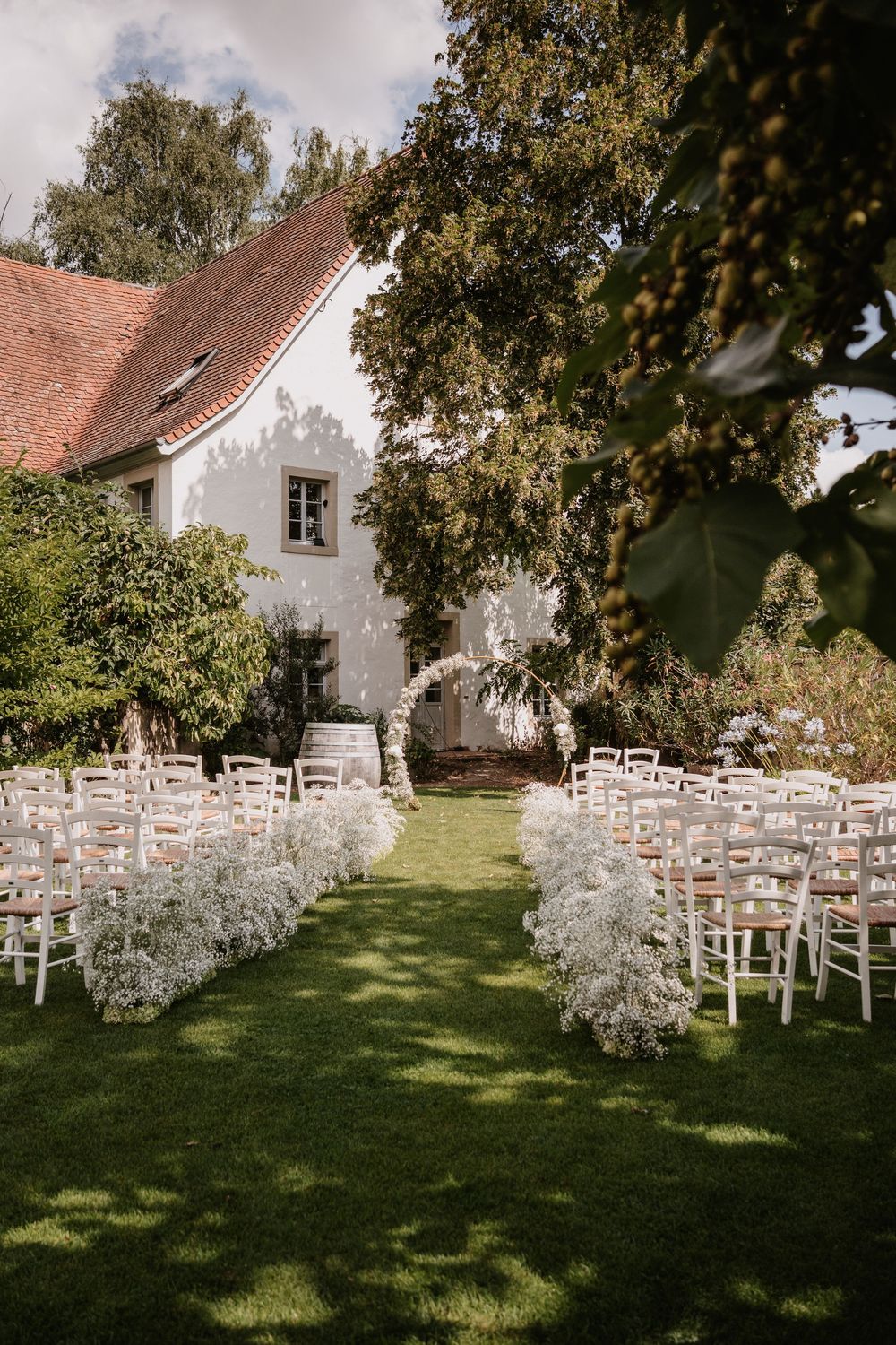 White wooden chairs arranged in rows on a lush green lawn for an outdoor wedding ceremony beside a rustic white building.