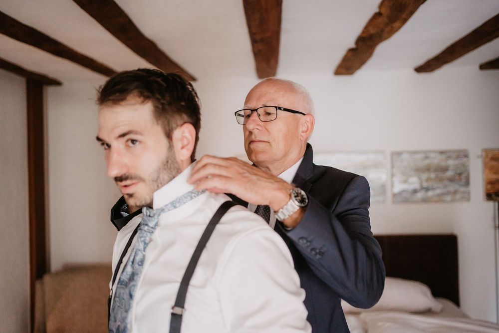 Groomsmen help adjust a bowtie and suspenders while getting ready for a wedding in a rustic room with wooden beams.