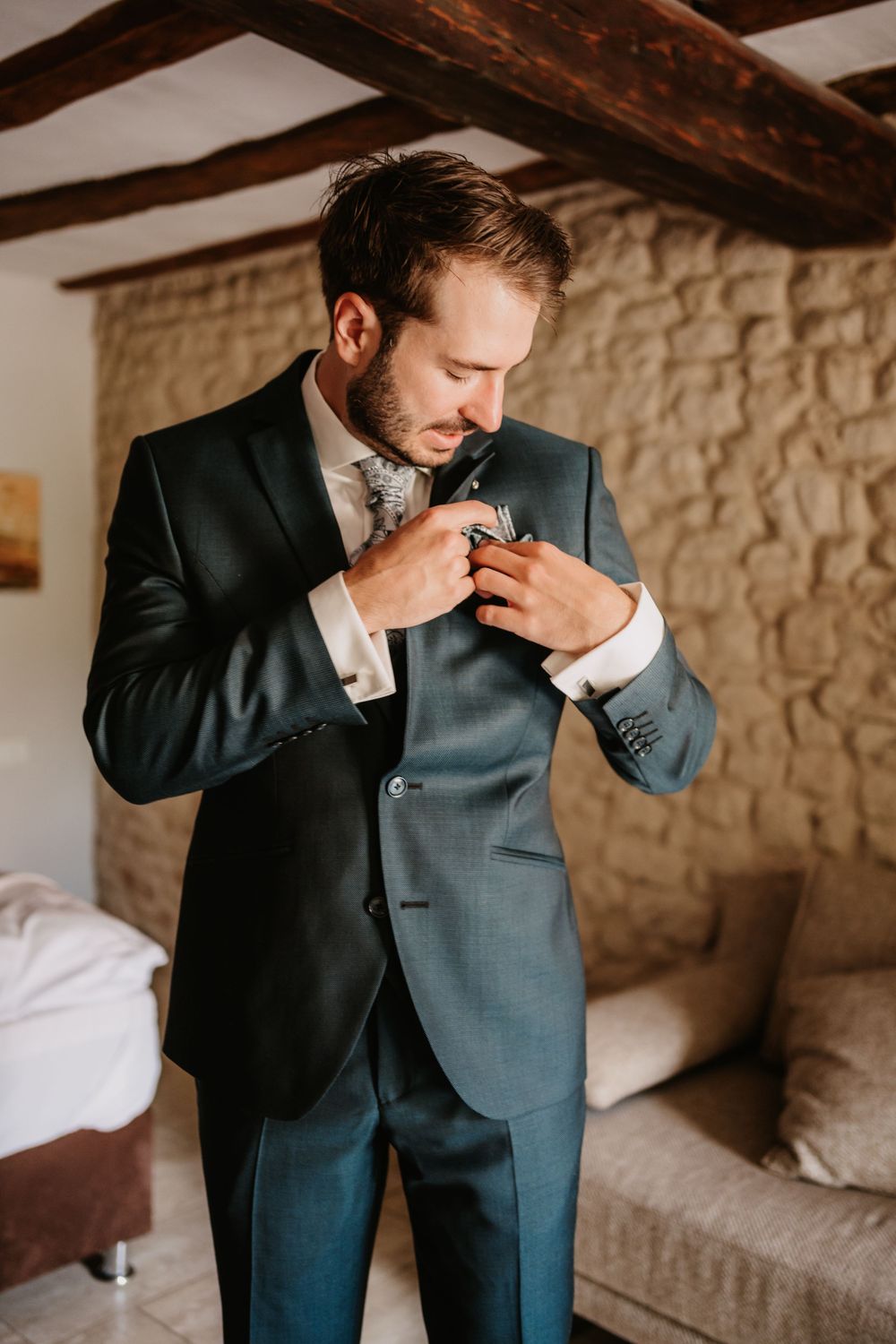 Groom in navy suit adjusts his tie while getting ready in rustic stone-walled room with wooden beams.