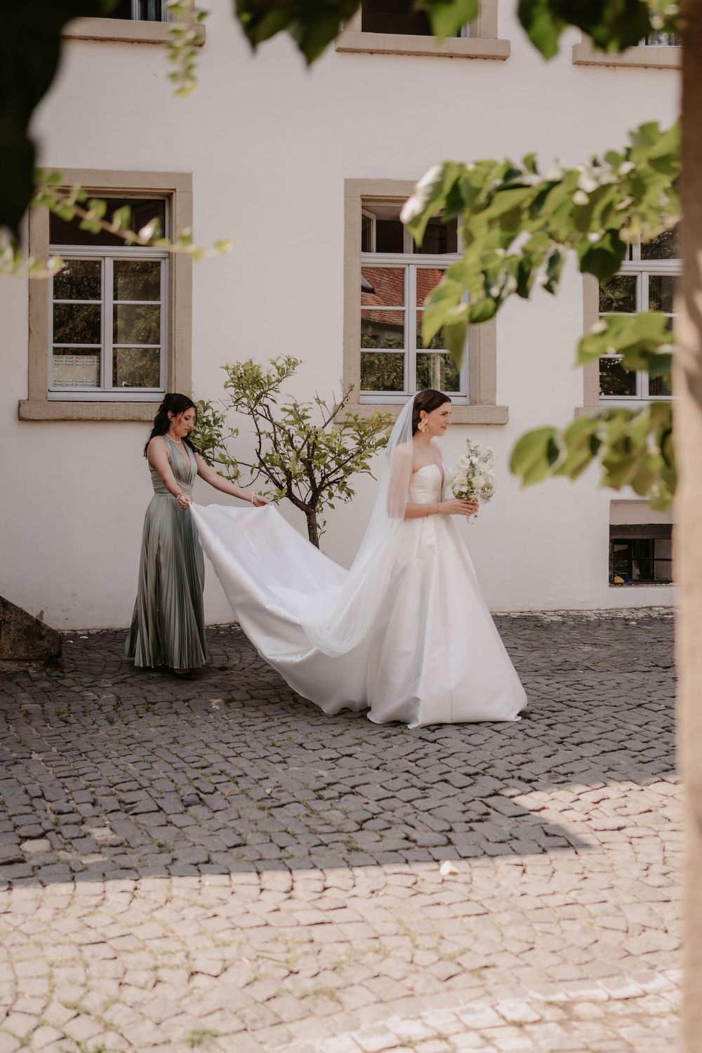 A bride in a flowing white gown walks on a cobblestone courtyard while her dress train is carried behind her.