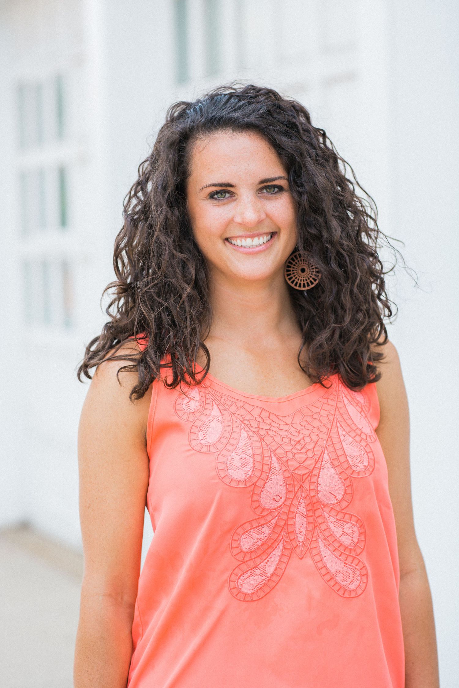 Woman in peach colored top with curly dark hair smiling against white wall.