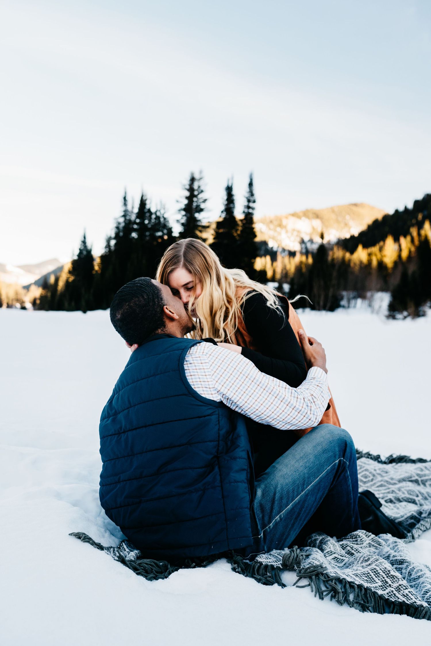 Couple snuggling together on snowy ground with evergreen trees and sunlit mountains in winter landscape.