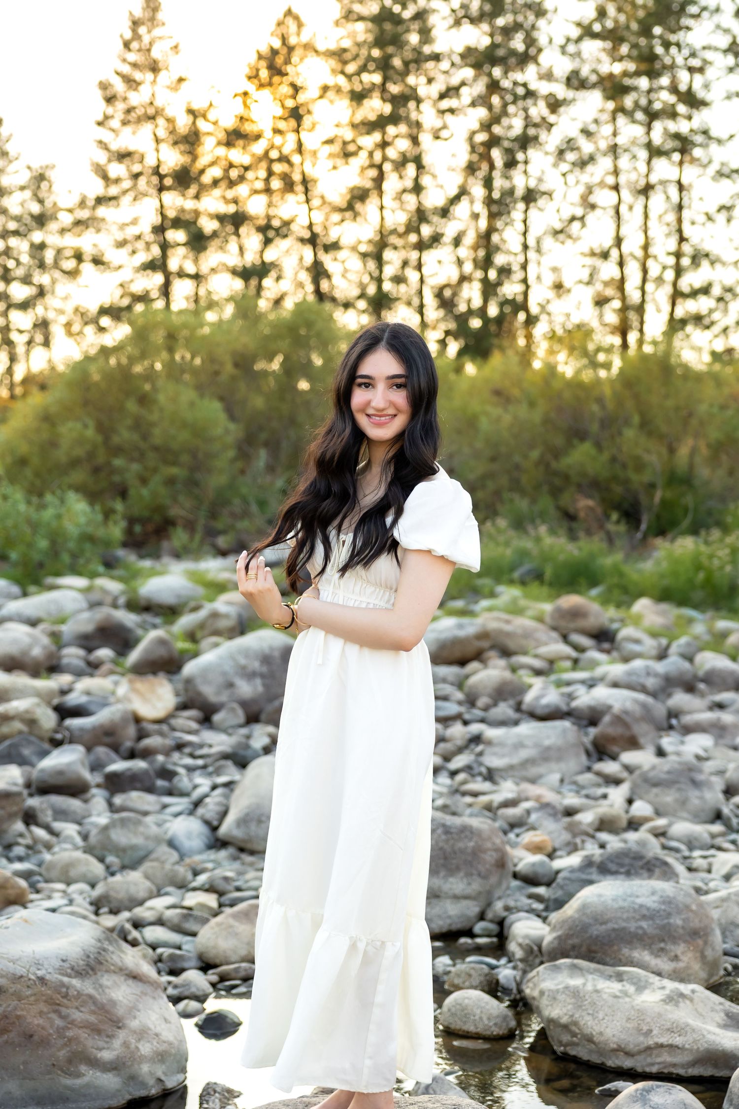 Person in a flowing white dress poses gracefully among rocky terrain at sunset with tall pine trees in the background.