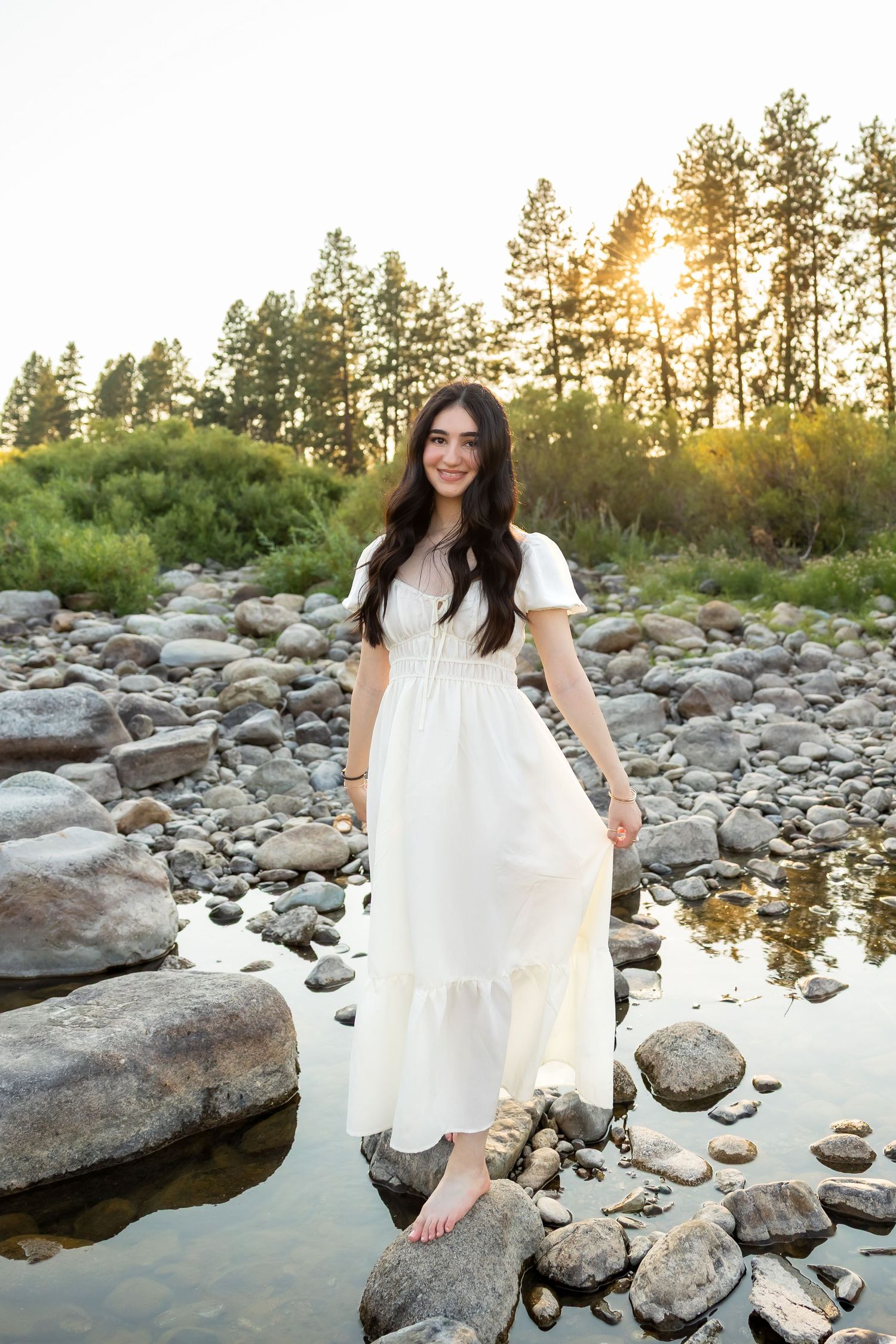 A white dress flows elegantly against rocky riverbank scenery with evergreen trees and golden sunlight.