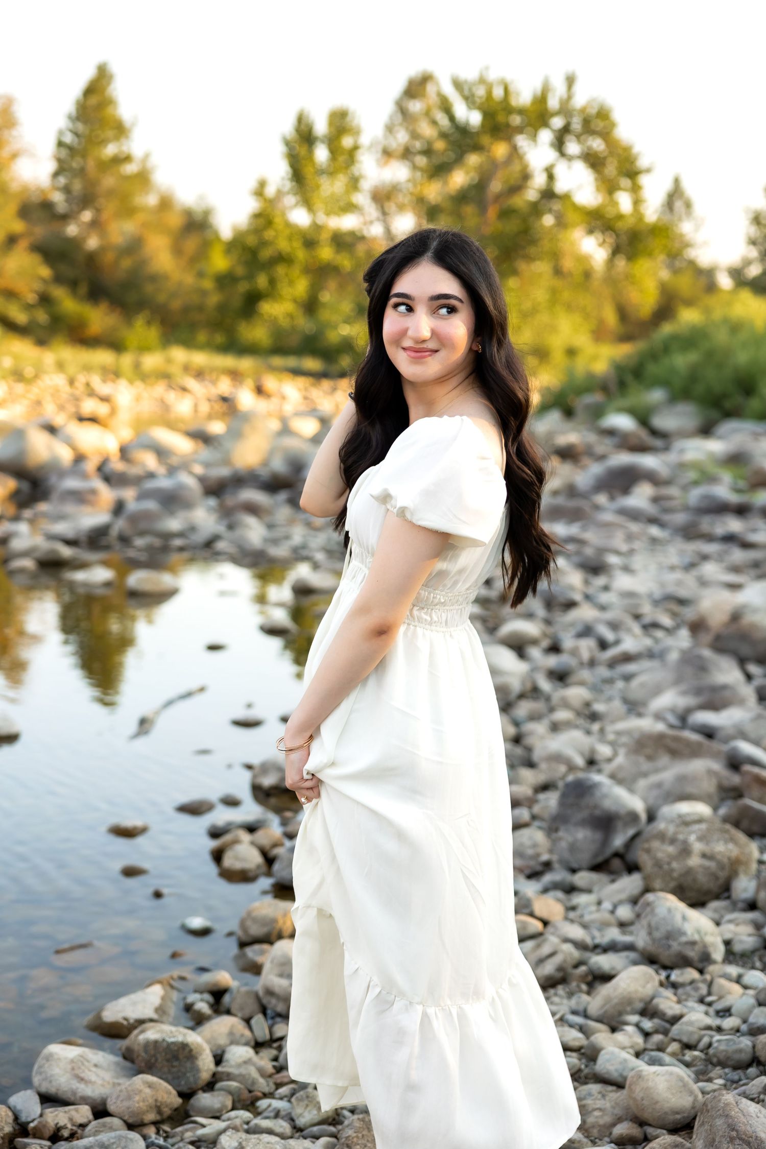 A white dress flows gracefully as someone poses by a rocky riverbank during golden hour.
