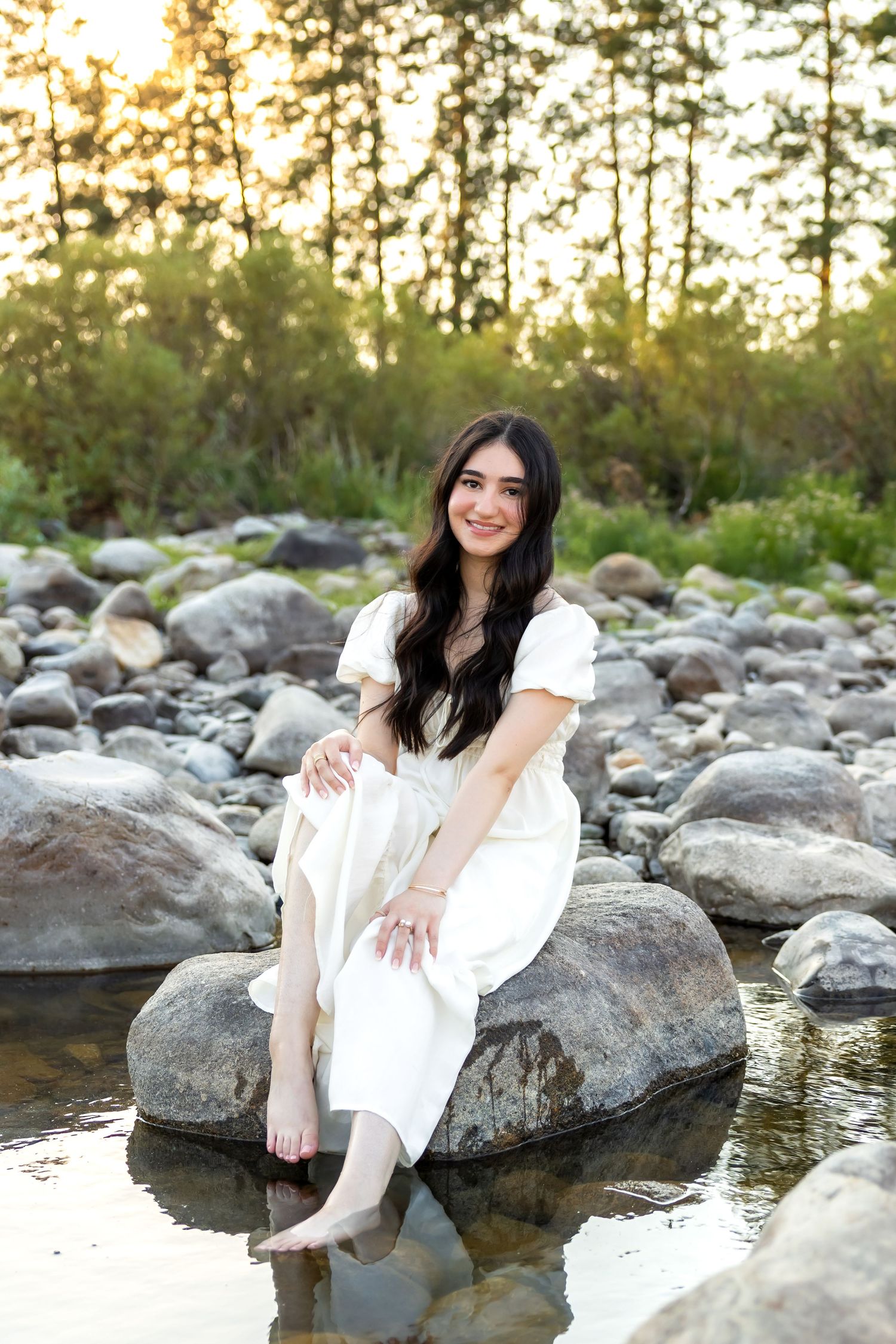 A person in a white dress poses on rocky terrain near a stream at sunset.