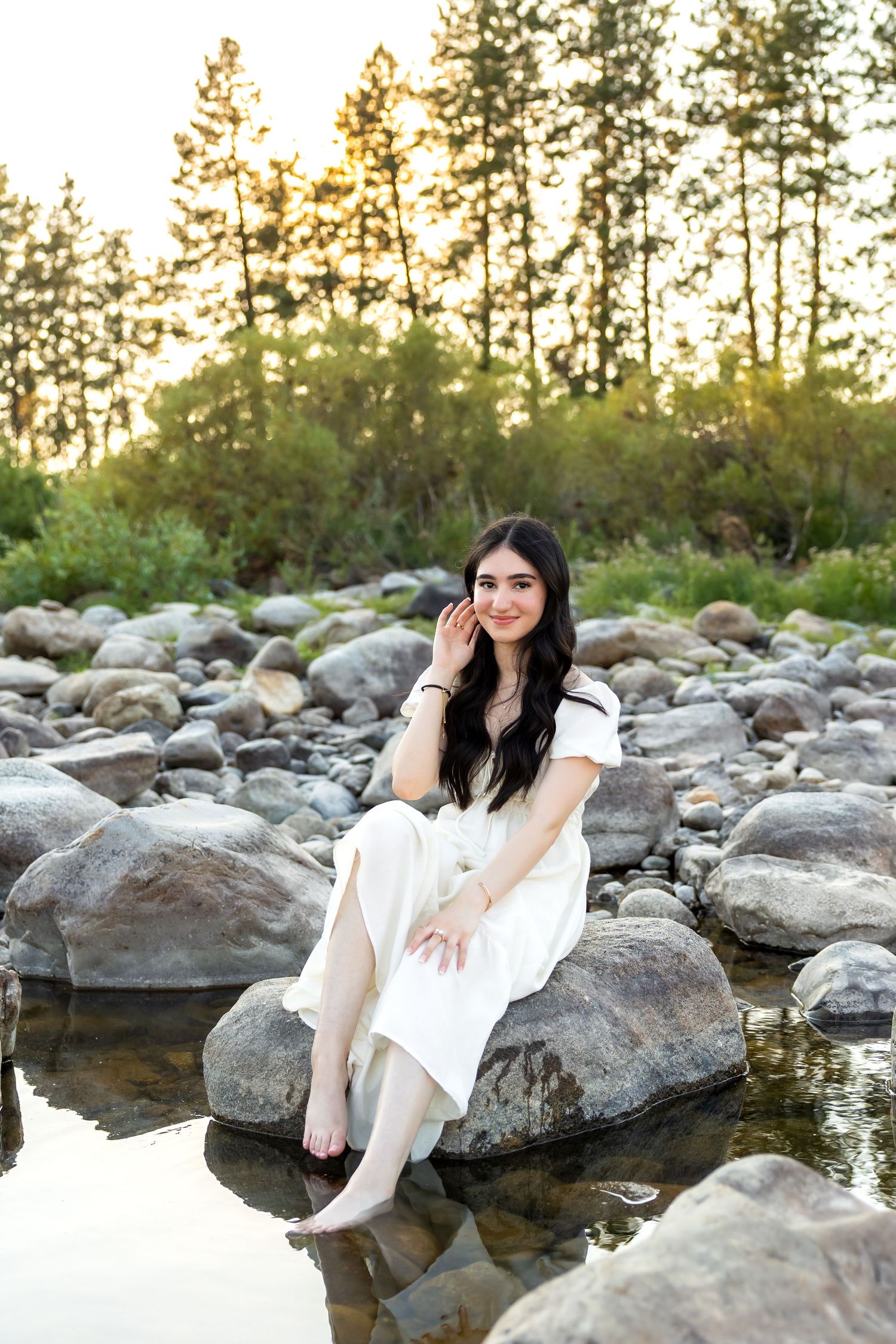 Someone in a white dress sits peacefully on rocks by a reflective stream with tall pines in background.